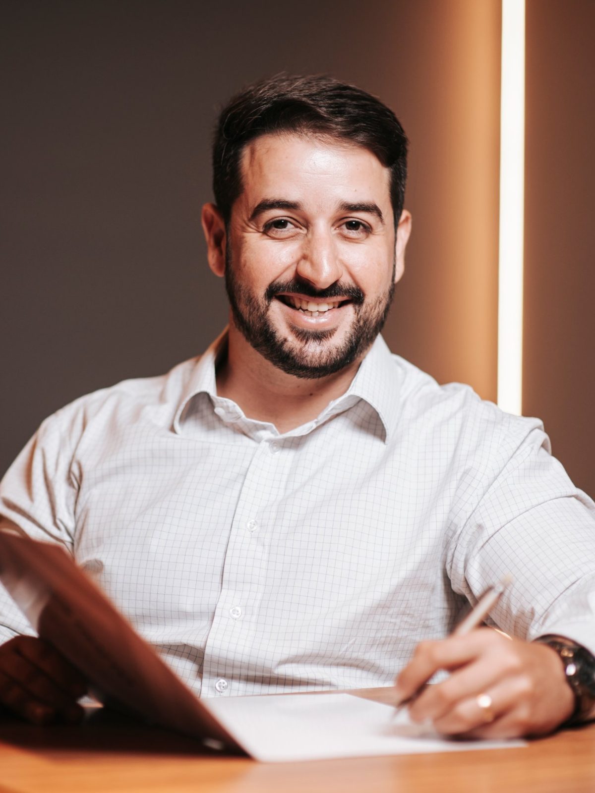 Retrato corporativo de homem de barba sorrindo à mesa, com papéis e caneta